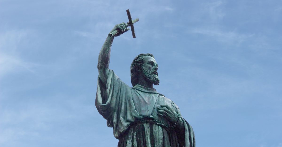 Estatua de bronce de Pedro El Ermitaño sosteniendo un crucifijo por encima de su cabeza y en el fondo de la imagen se ve el cielo azul despejado