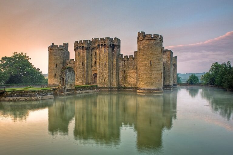 Imagen del castillo de Bodiam con una puesta del sol en el bondo de la imagen. Se ve el reflejo del castillo sobre el agua que rodea el castillo