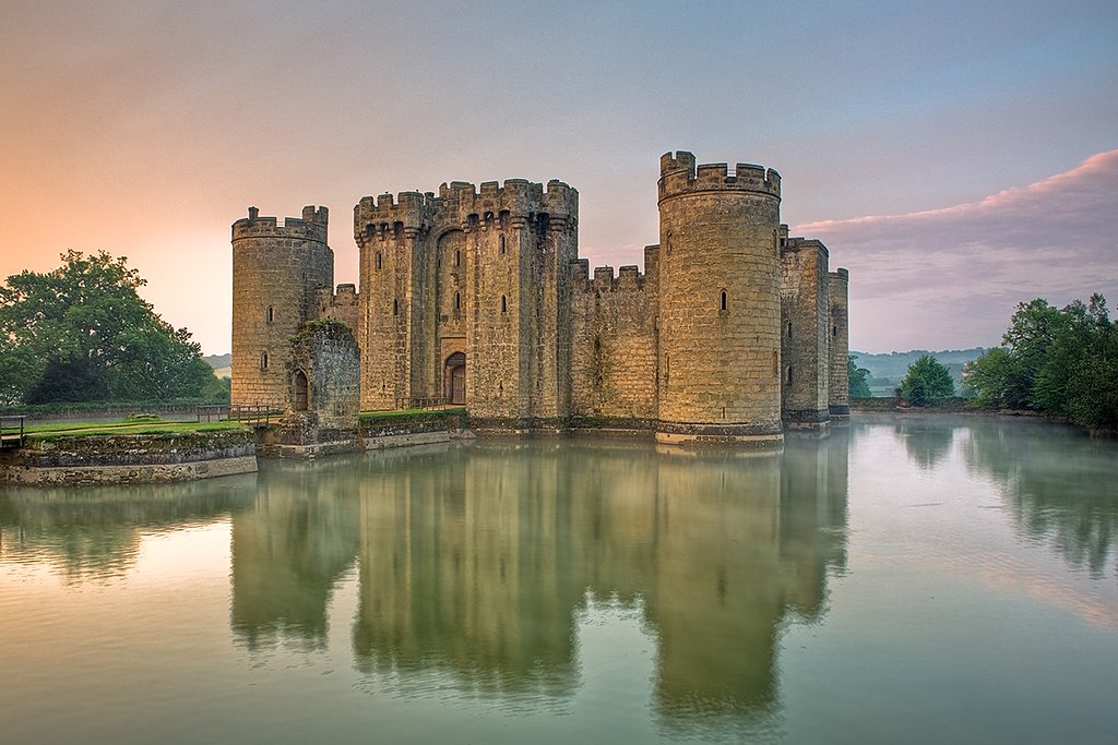 Imagen del castillo de Bodiam con una puesta del sol en el bondo de la imagen. Se ve el reflejo del castillo sobre el agua que rodea el castillo